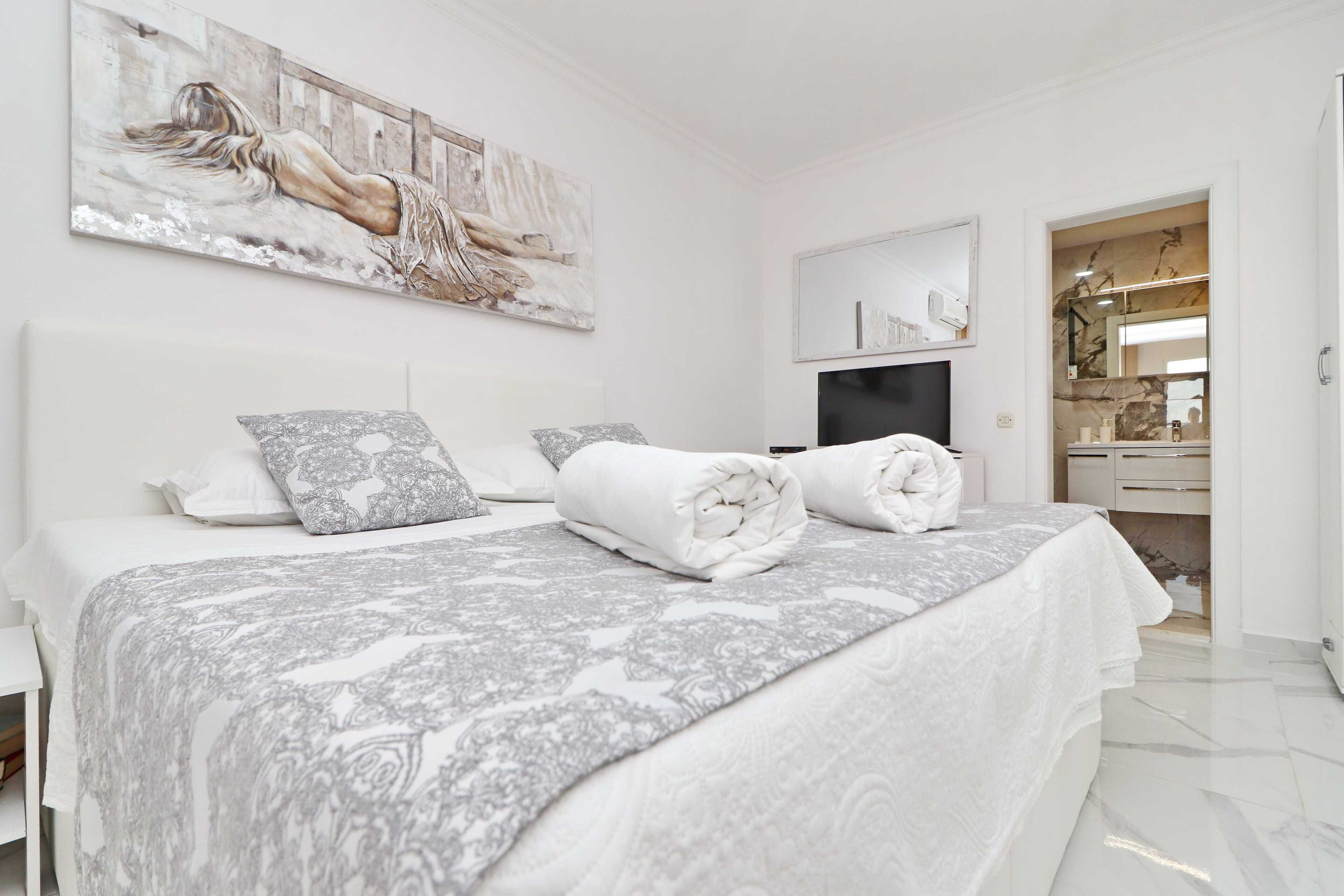 A modern bedroom with a large bed, a painting on the wall, and a bathroom visible through an open door.