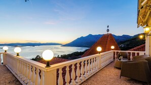 Un balcon doté d’une balustrade blanche, une table avec des chaises, et une vue sur une montagne et une étendue d’eau au coucher du soleil.