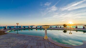 Un espace piscine agrémenté de chaises longues et d’un bac à fleurs décoratif, offrant une vue sur un plan d’eau et un coucher de soleil.