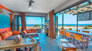 A modern living room with a large window overlooking the beach, a sofa with orange cushions, a dining area with wooden tables and chairs, and a ceiling fan. - Skinos ilivatos Boutique Eco Villa (Kefalonia)