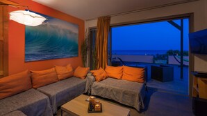 A modern living room with a large ocean wave painting, a sofa with orange cushions, and a view of the sea through a large window. - Skinos ilivatos Boutique Eco Villa (Kefalonia)
