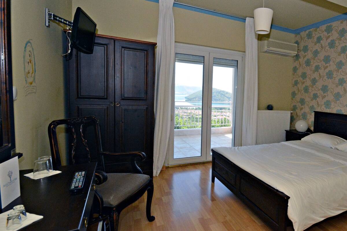 Een slaapkamer met een bed, een stoel, een bureau met een afstandsbediening, een nachtkastje met een lamp en uitzicht op een balkon met een stadsbeeld.