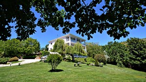 A white two-story house with a balcony, surrounded by lush greenery and a well-maintained lawn.