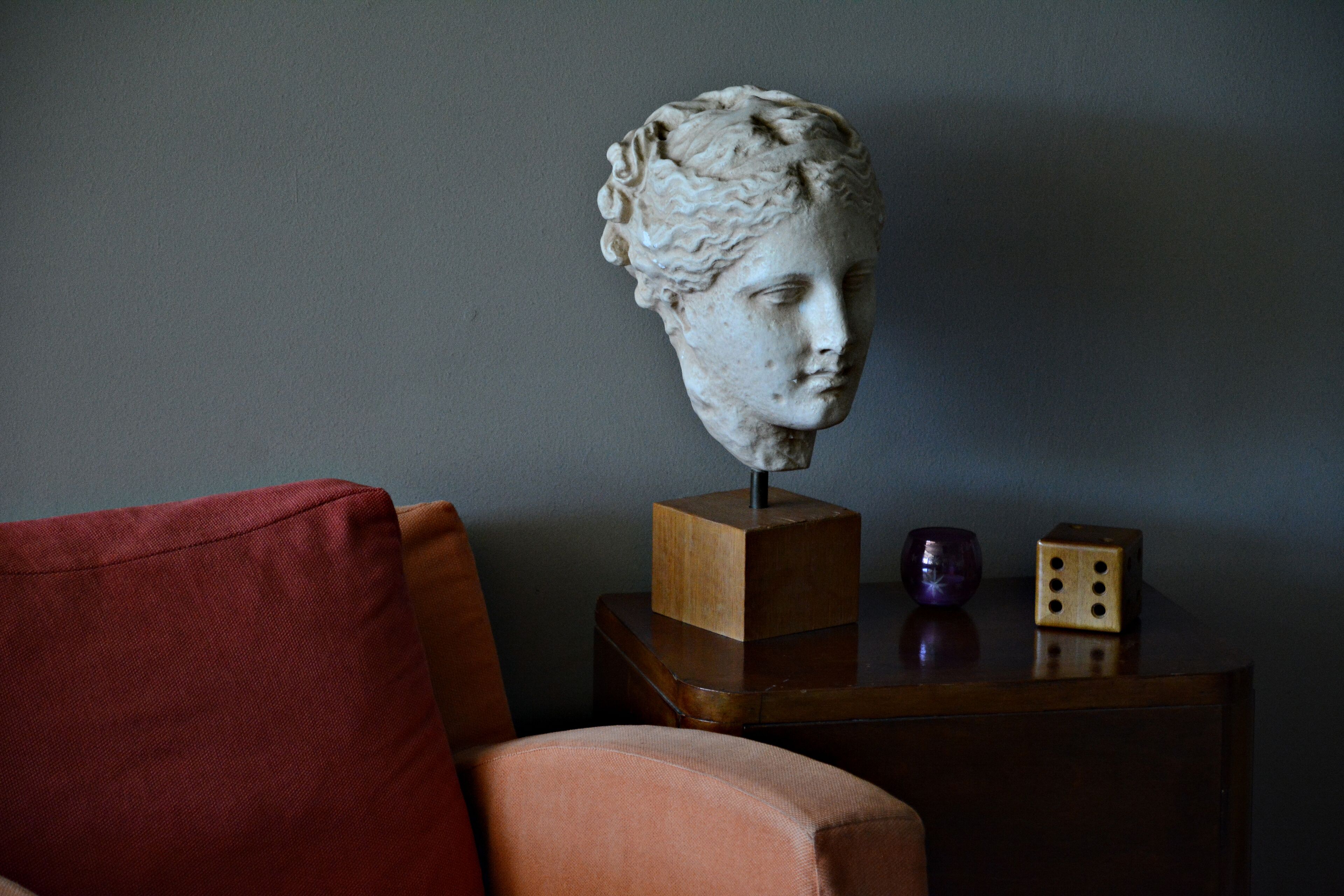 A bust sculpture on a wooden stand, a red cushion, and a small purple object on a table.