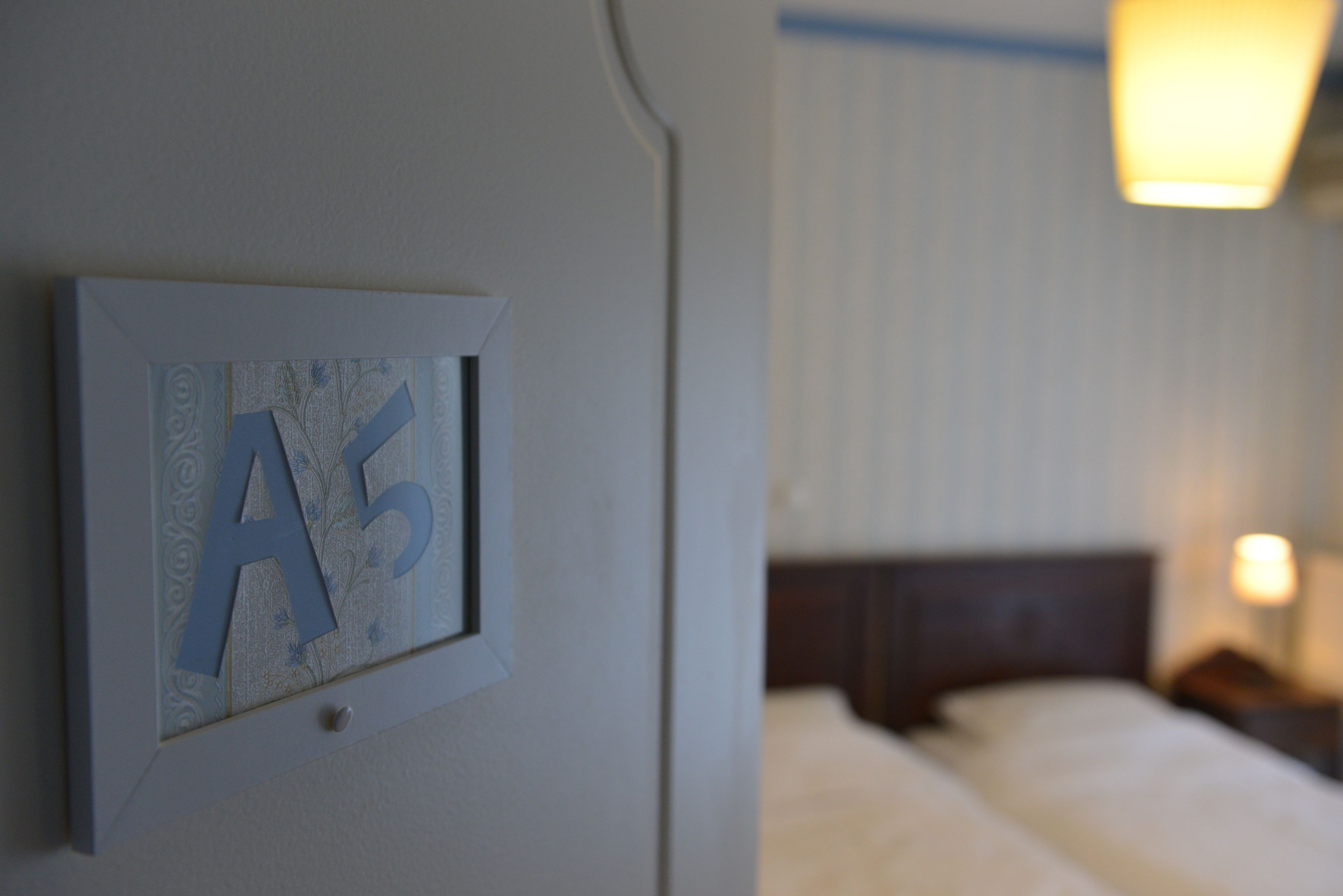 Et indrammet skilt med bogstaverne "A5" på en væg, en seng med hvide sengetæpper og et natbord med en lampe i et hotelværelse.