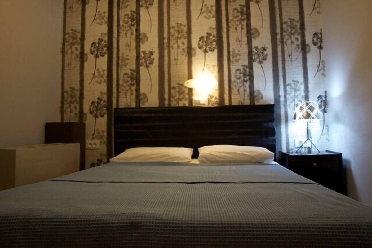 A neatly made bed with a dark headboard, a nightstand with a lamp, and a wall with floral wallpaper.