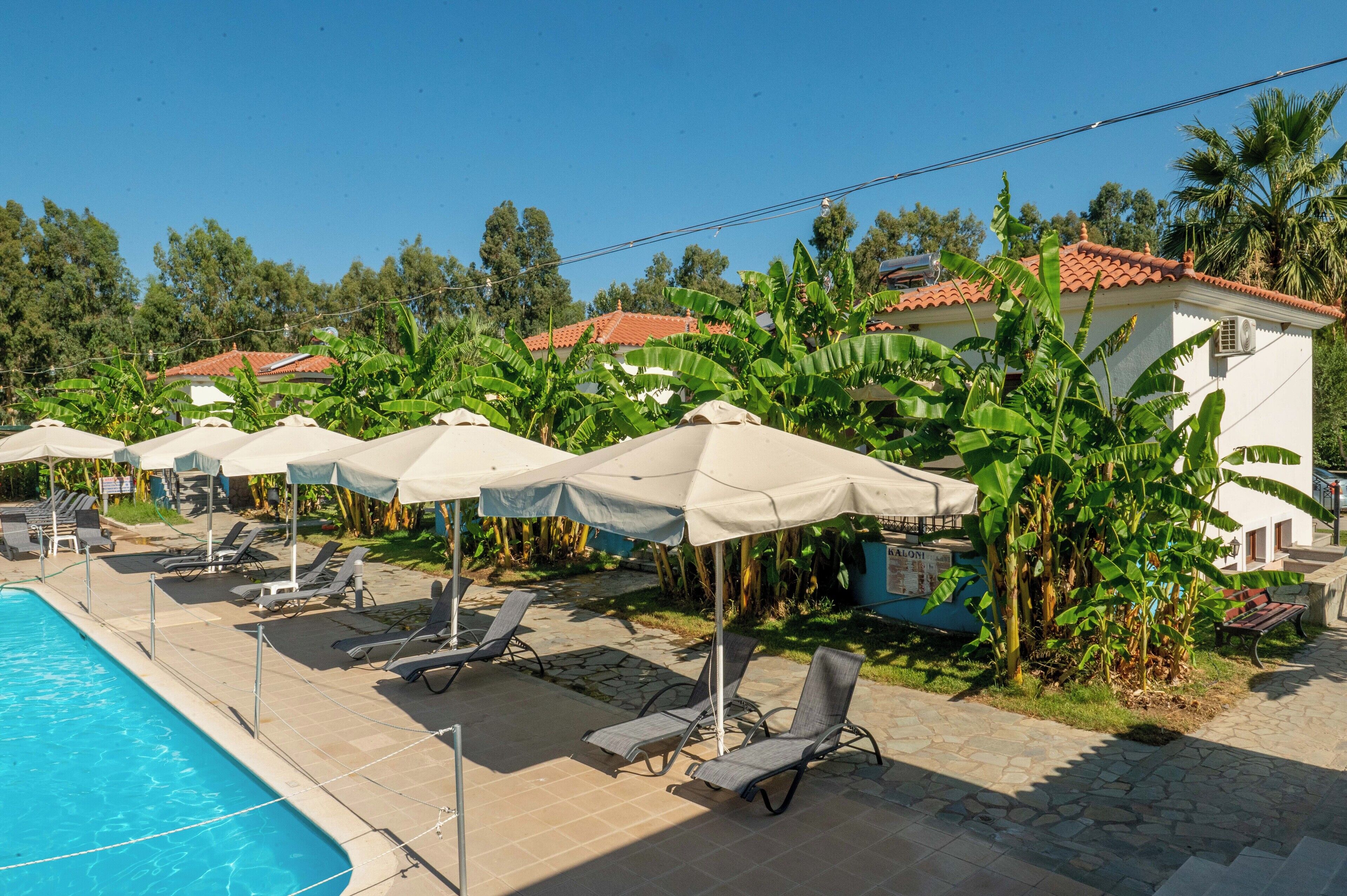 A poolside area with lounge chairs and umbrellas, surrounded by banana trees and a building with a red-tiled roof.