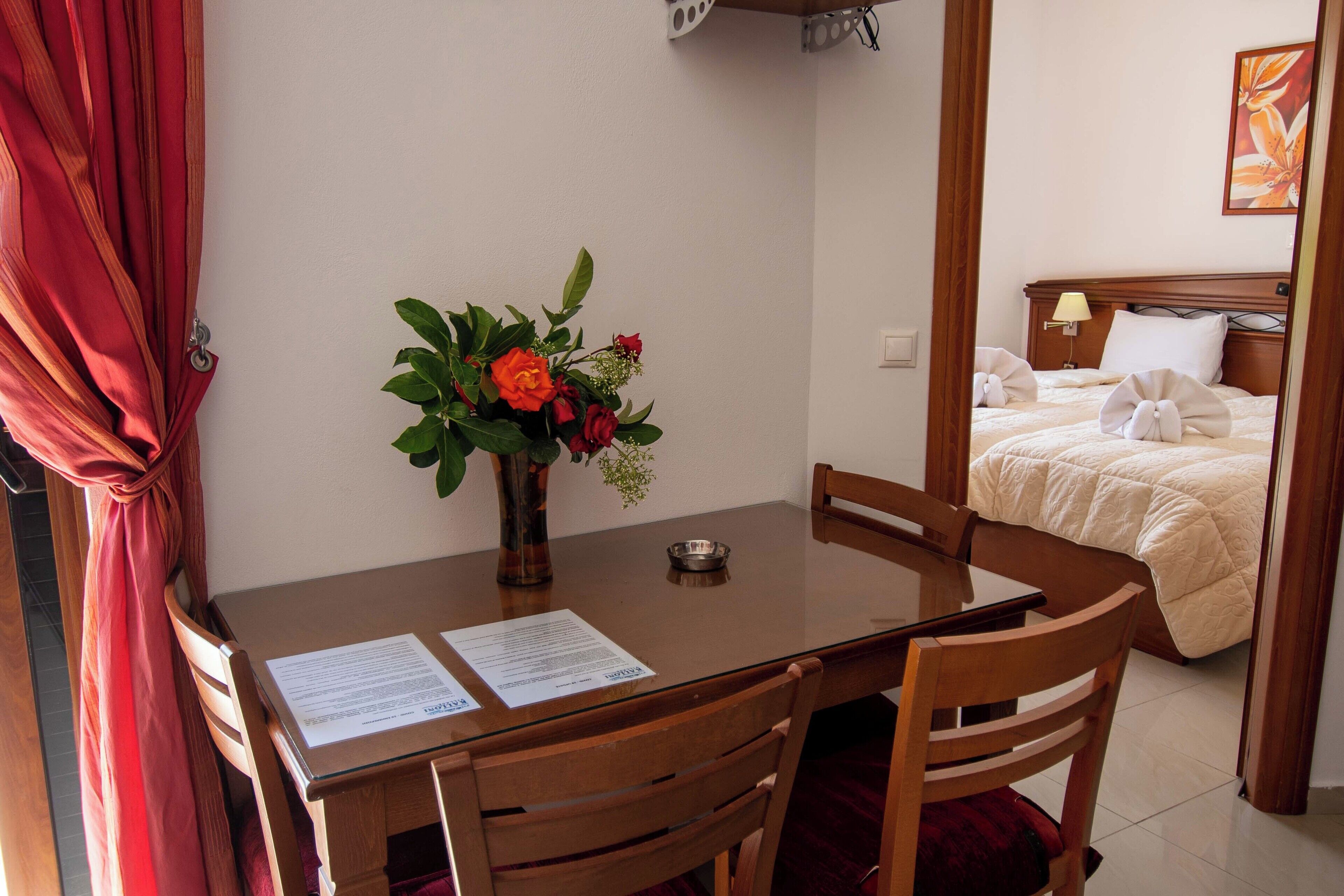 A hotel room with a wooden dining table, chairs, a vase with flowers, a bed with a white comforter, and a painting on the wall.
