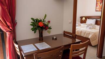 A hotel room with a wooden dining table, chairs, a vase with flowers, a bed with a white comforter, and a painting on the wall.