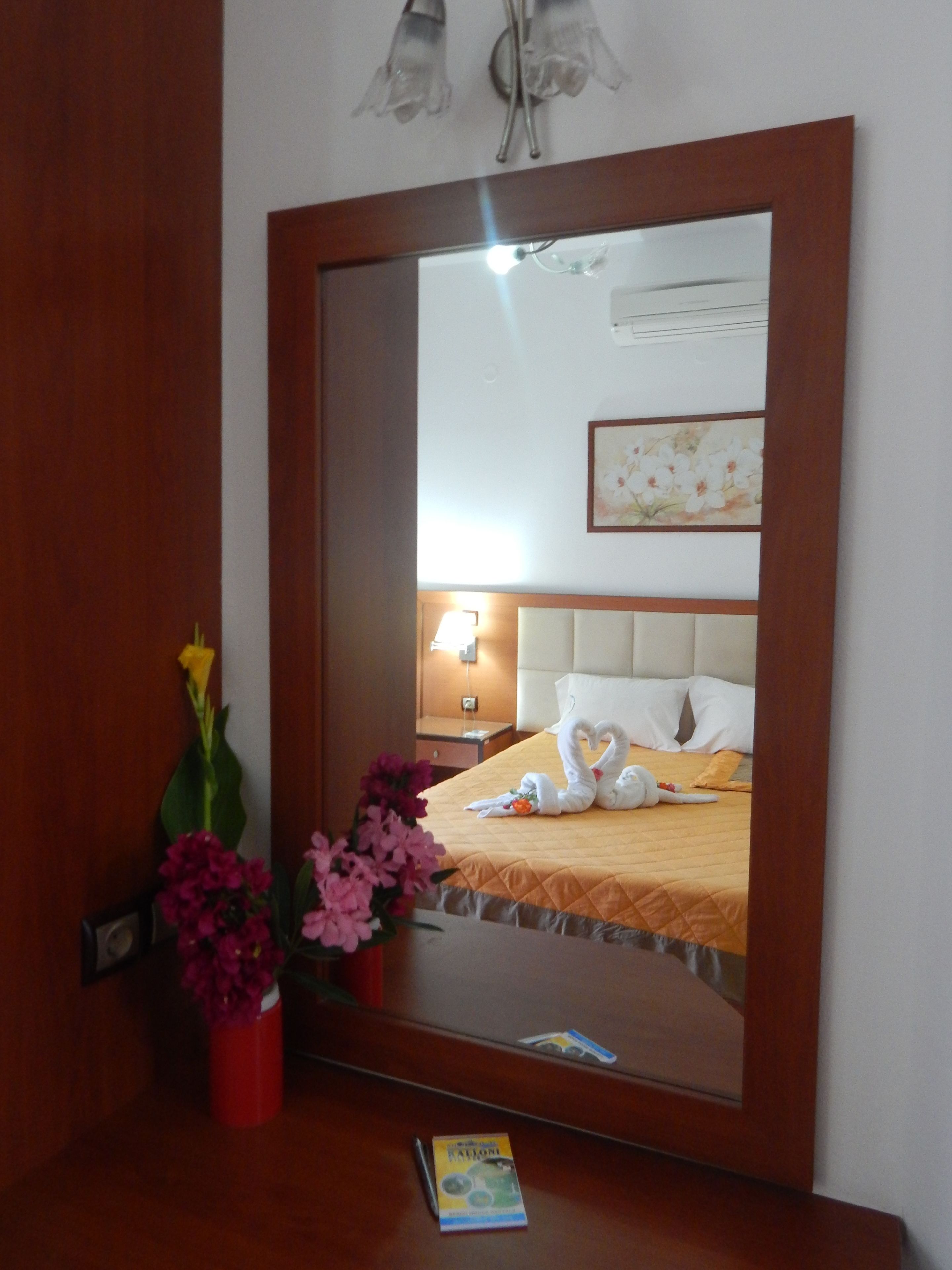 A hotel room with a large mirror, a bed with a white headboard, a bedside table with a lamp, and a vase with flowers.