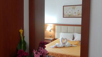 A hotel room with a large mirror, a bed with a white headboard, a bedside table with a lamp, and a vase with flowers.