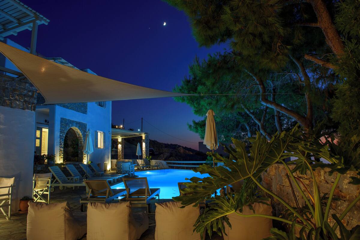 A pool area with lounge chairs and umbrellas, a stone wall, and a view of the night sky.