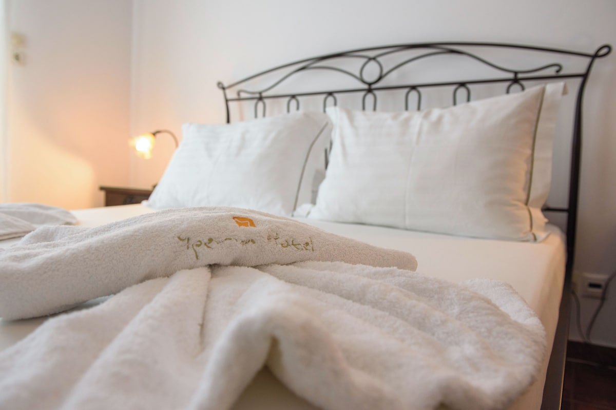 Un lit bien fait, avec du linge de lit blanc et une serviette ornée d’un motif, une tête de lit en métal et une lampe de chevet.