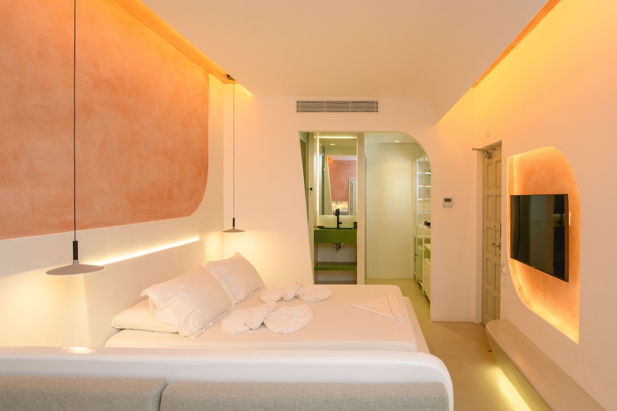 A modern hotel room with a large bed, bedside lamps, a TV, and a bathroom visible through an open door.