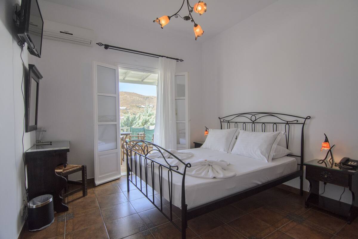 A bedroom with a metal bed frame, white linens, a bedside table with a lamp, a small chair, and a view of the outdoors through a window.