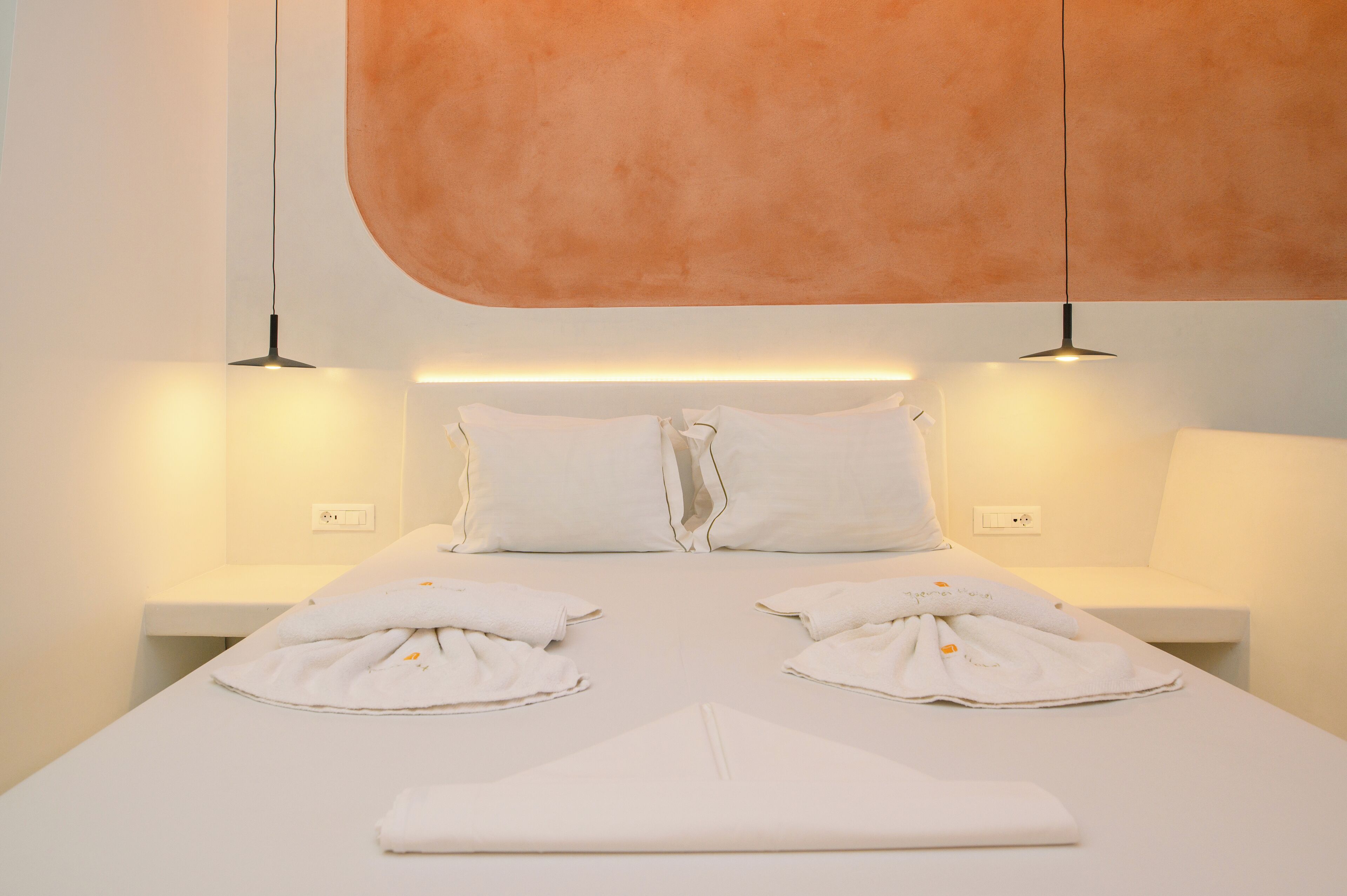 A neatly made bed with pillows and towels, a white bedside table, and a wall-mounted light fixture.