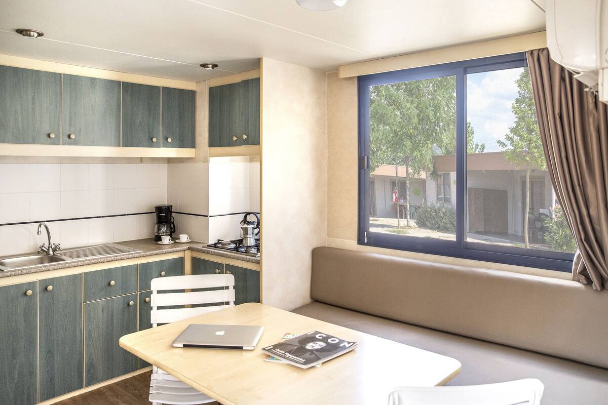 A compact kitchen with a dining area, a window with curtains, and a laptop on the table.
