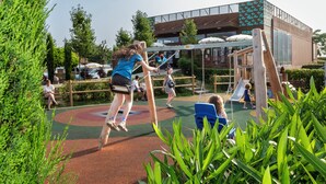 Ein Spielplatz mit Schaukeln und Rutschen, umgeben von viel Grün und einem Gebäude mit großen Fenstern.