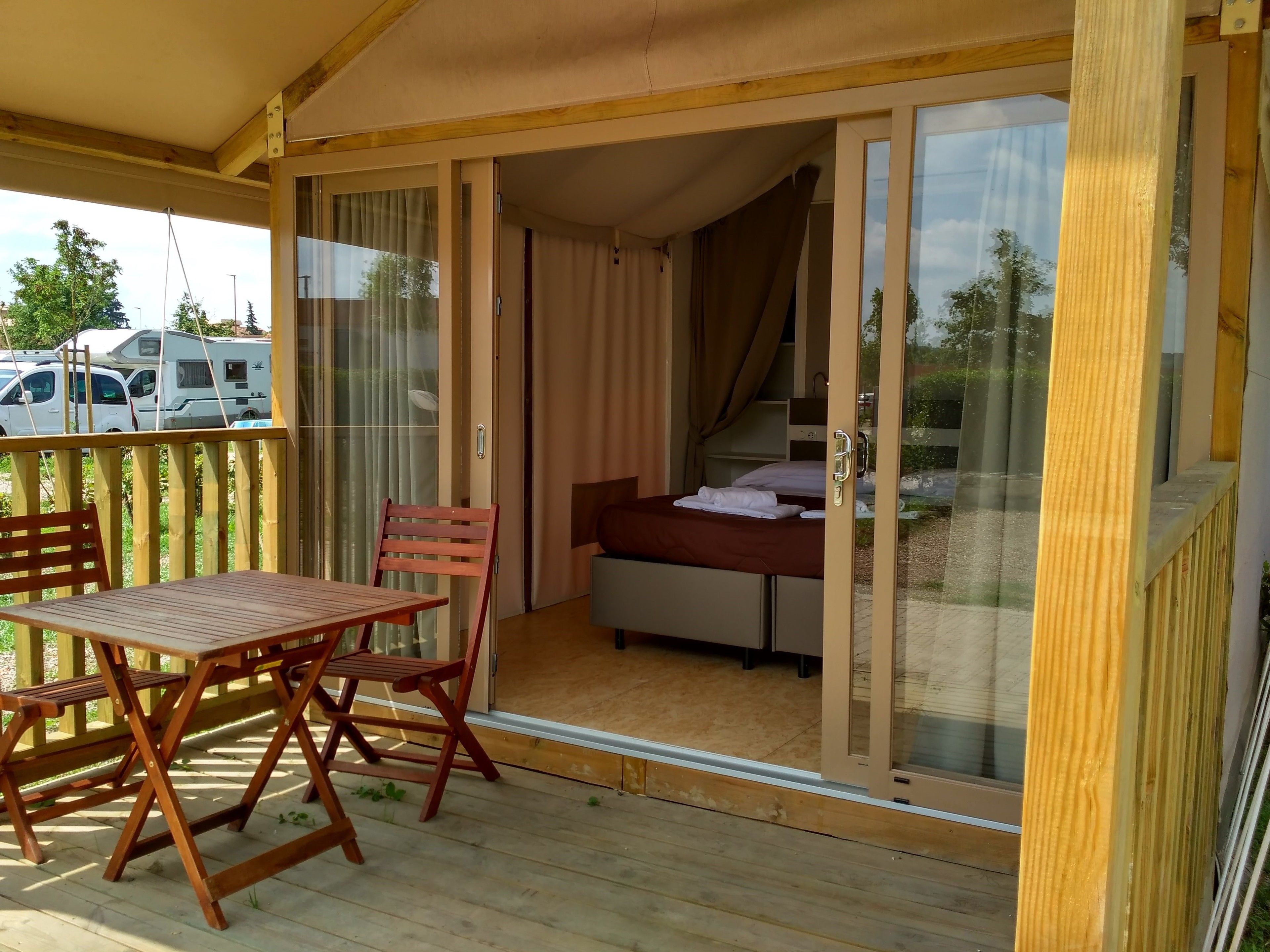 Een balkon met een houten tafel en stoelen, een bed met een witte matras en een bruin hoofdeinde, en een schuifdeur die naar een kamer leidt.