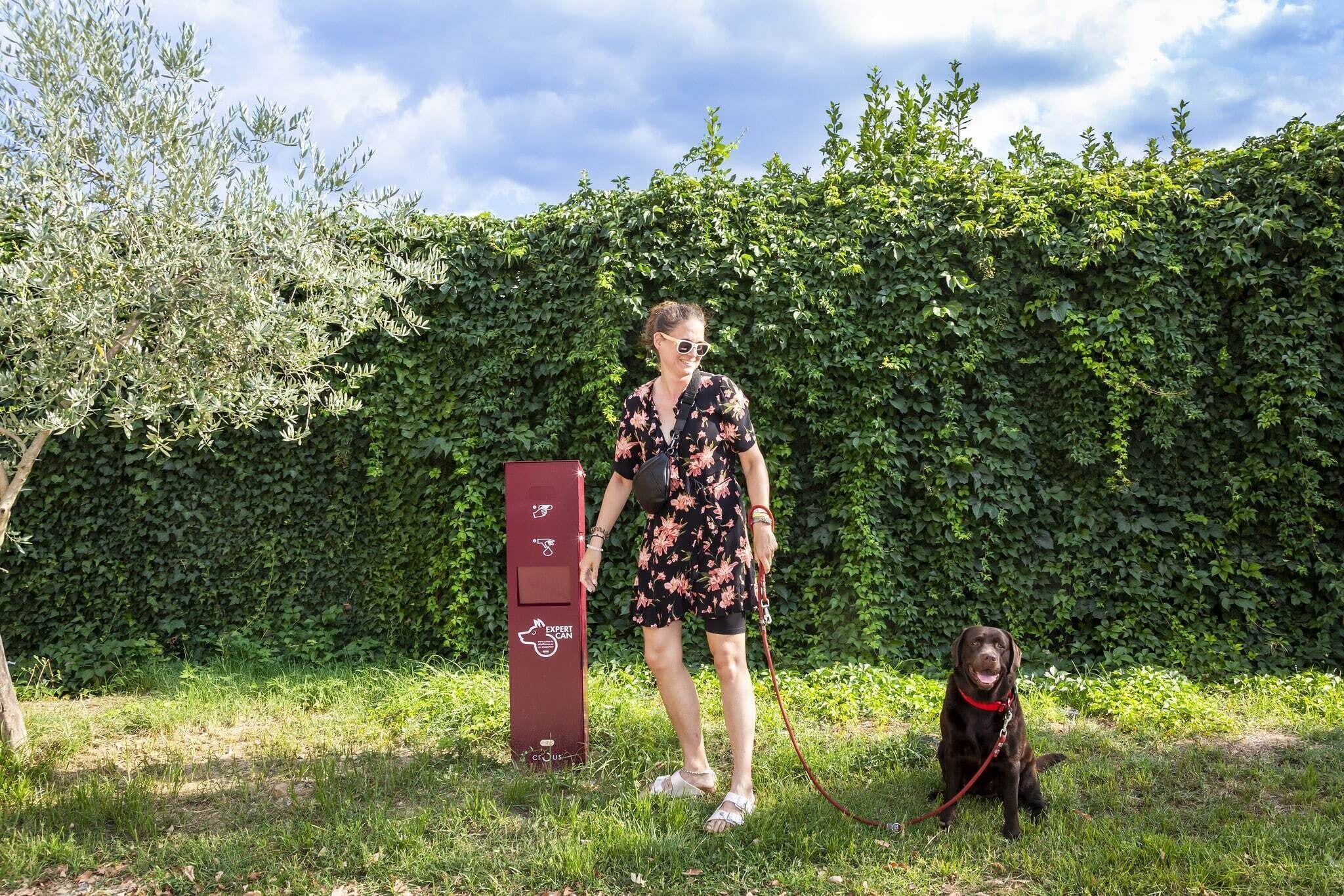 A person with a dog in a grassy area with a hedge in the background and a red electric vehicle charging station.