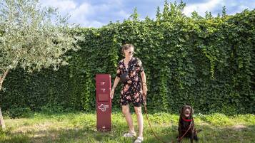 A person with a dog in a grassy area with a hedge in the background and a red electric vehicle charging station.