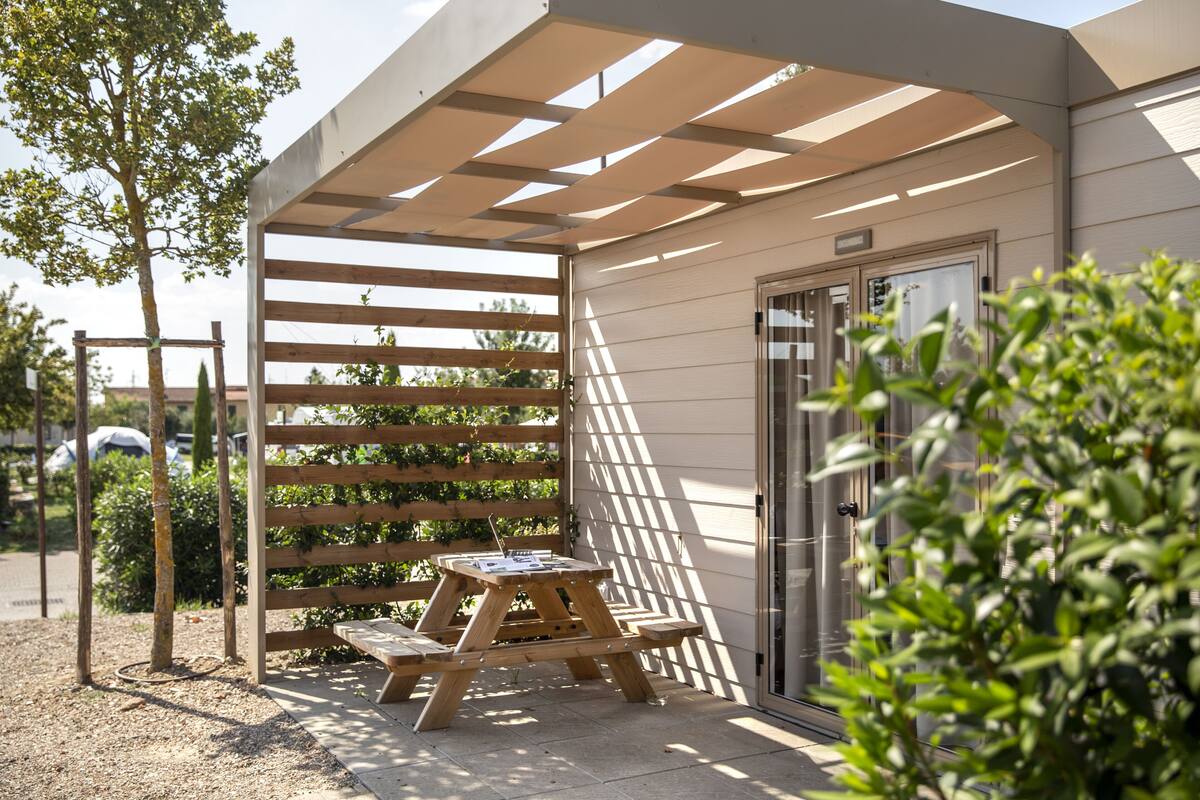 A covered patio area with a wooden table and benches, a pergola providing shade, and a door to the building.