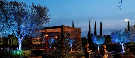 Un espacio exterior con mesas, un edificio moderno, árboles y un banco.