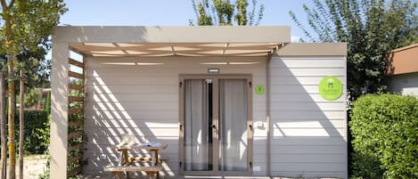 A small, single-room building with a door and a window, a wooden bench, and a sign with a logo.