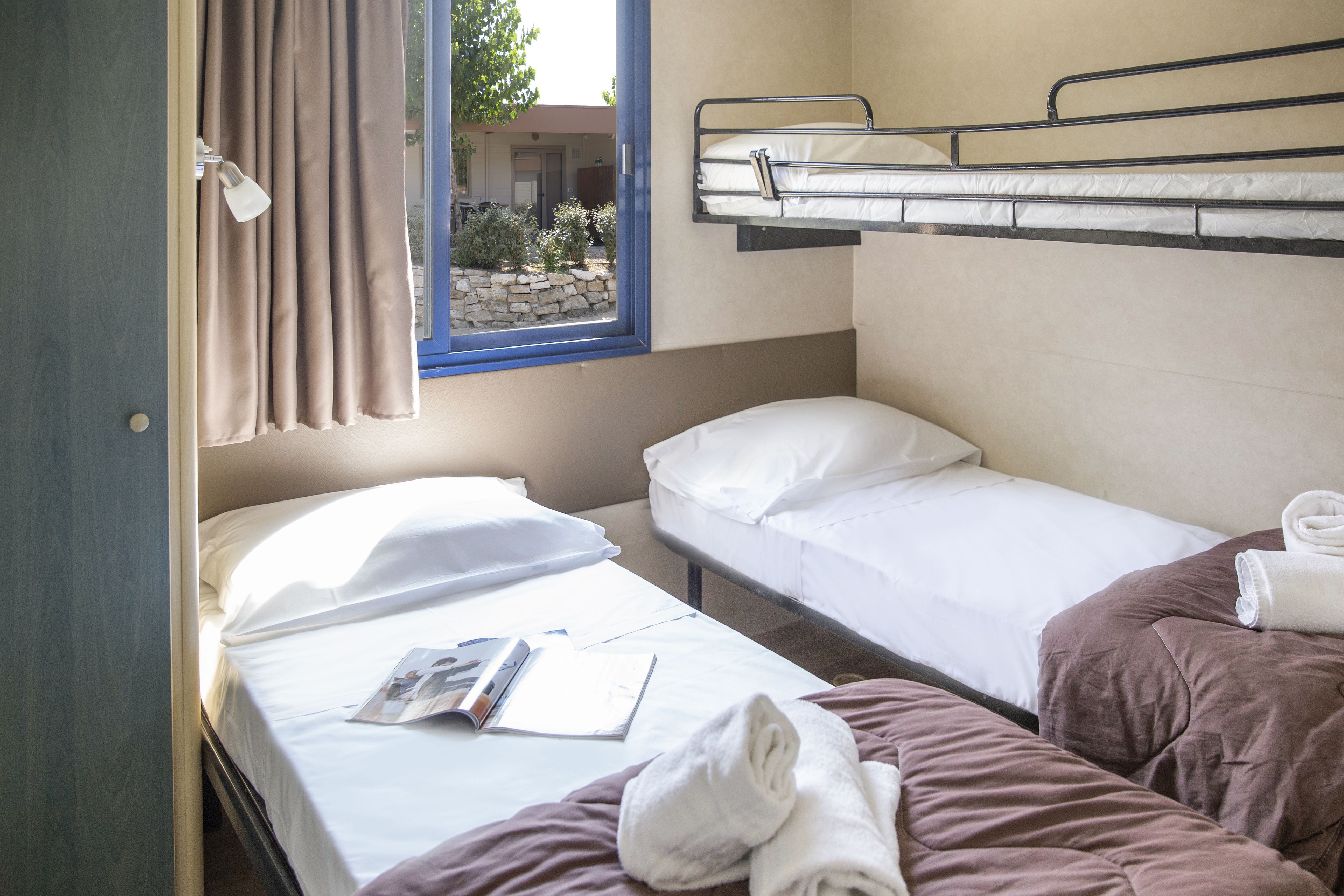 A small, compact room with two beds, one with a window and curtains, and a bunk bed above.