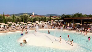 Ein großer Pool im Freien mit mehreren Wasserspielen, umgeben von Liegestühlen und Sonnenschirmen, mit Blick auf Hügel im Hintergrund.