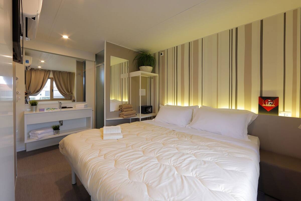 A hotel room with a large bed, a bedside table, a mirror, a vanity with a sink, and a potted plant.