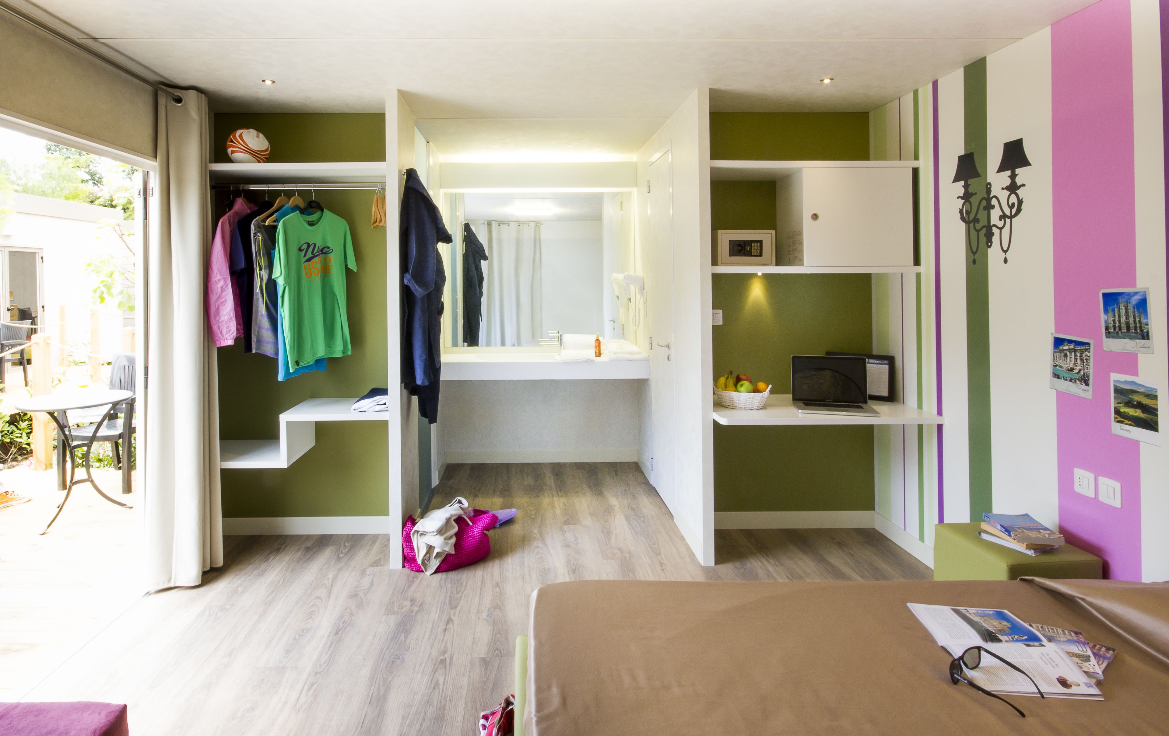 Una stanza con un letto, una scrivania e una sedia. Si vede una finestra con tende, un armadio pieno di vestiti e uno scaffale con libri e un computer portatile.