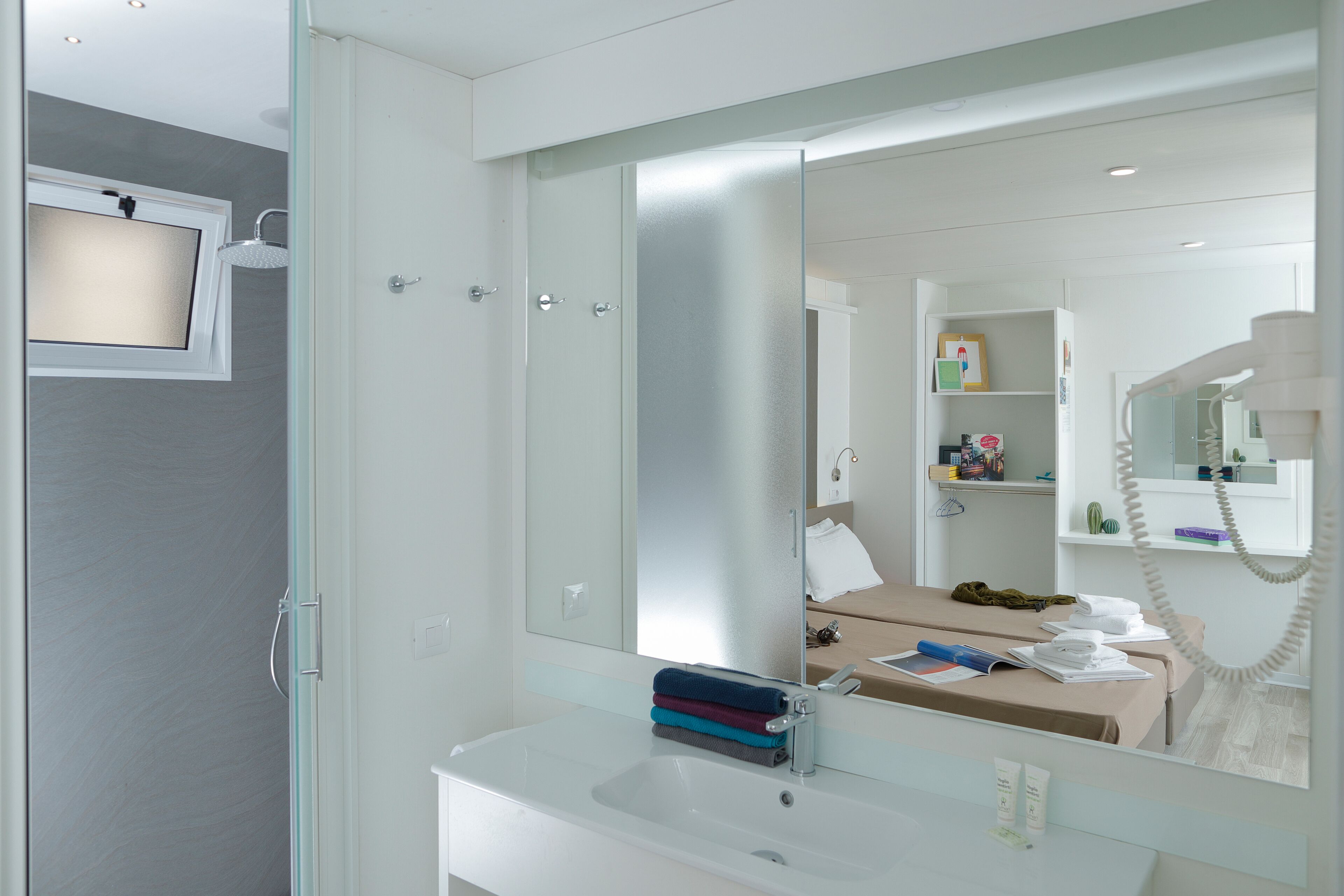 A modern bathroom with a glass shower enclosure, a white sink, and a large mirror reflecting a bed and a telephone.