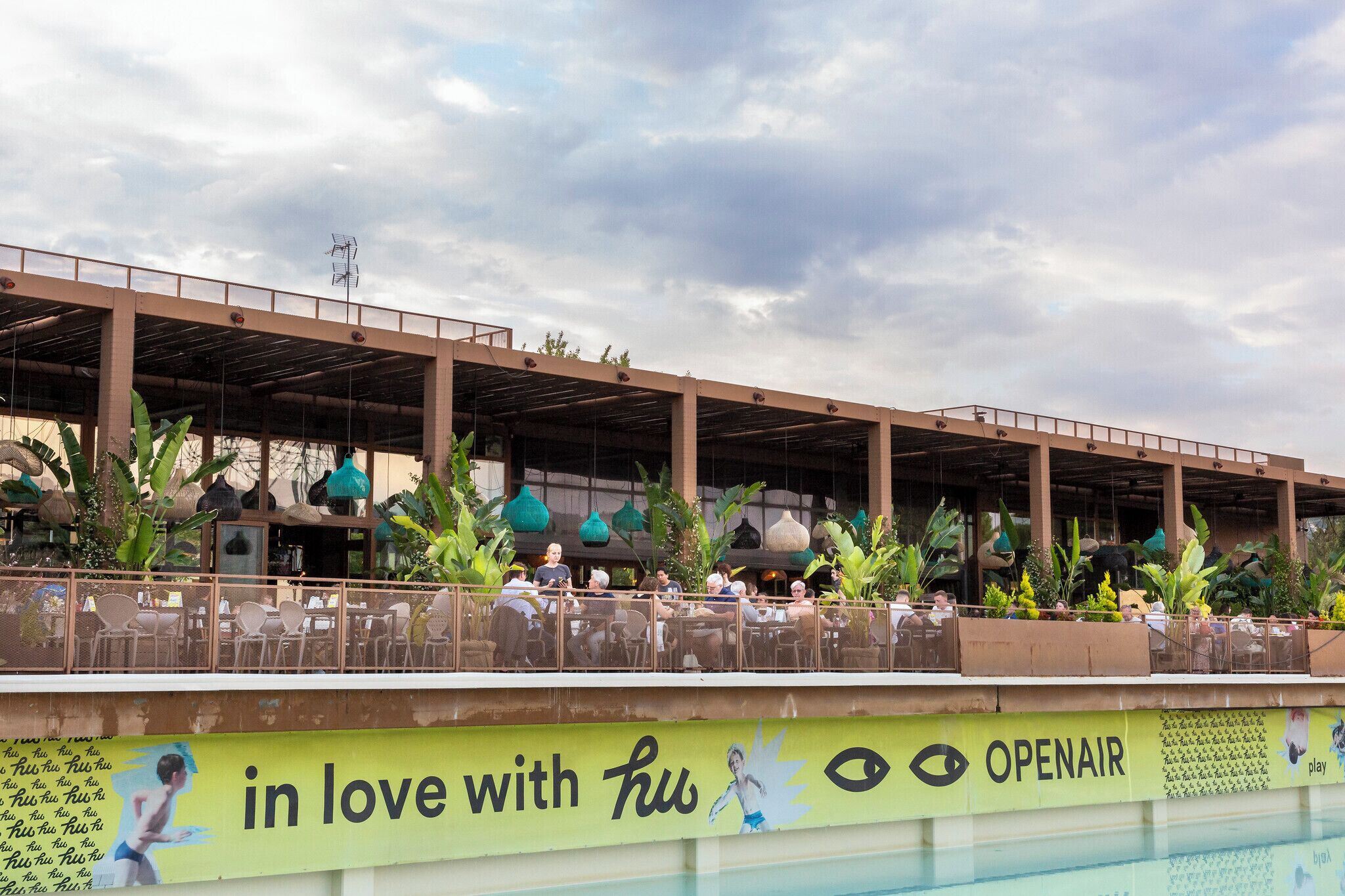 An outdoor restaurant with a covered seating area overlooking a pool, featuring a prominent OpenAir advertisement.