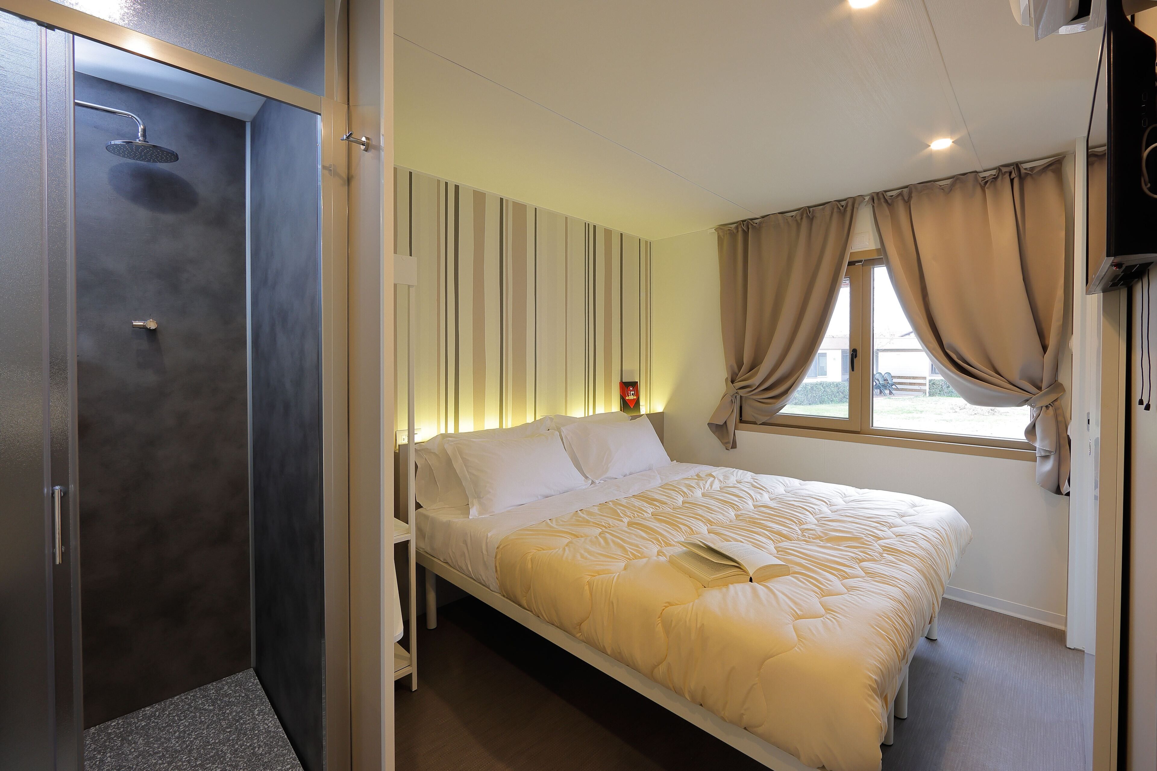 A bedroom with a bed, a shower, a window with curtains, and a wall-mounted TV.