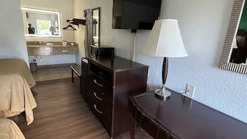 Une chambre d’hôtel avec un plancher en bois, un lit recouvert d’une couverture beige, un bureau avec une lampe, une commode, un téléviseur fixé au mur et une salle de bain visible au fond.