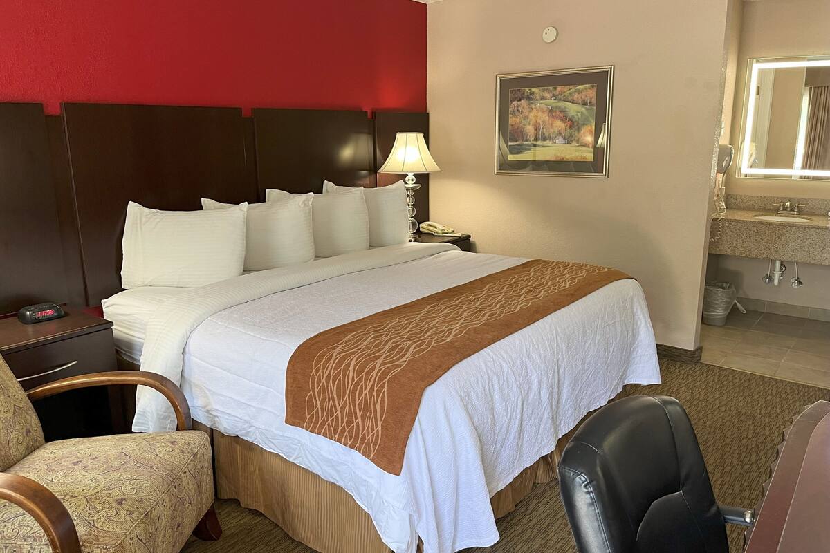Une chambre d’hôtel avec un grand lit, une table de chevet avec une lampe, une chaise et une salle de bain avec un lavabo et un miroir.