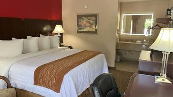 Habitación de hotel con una cama grande, un escritorio con lámpara, una silla, baño con lavamanos y espejo, y un cuadro en la pared.