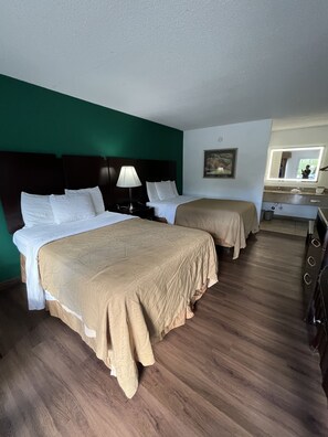 A hotel room with two beds, a nightstand with a lamp, a painting on the wall, and a bathroom visible through an open door.