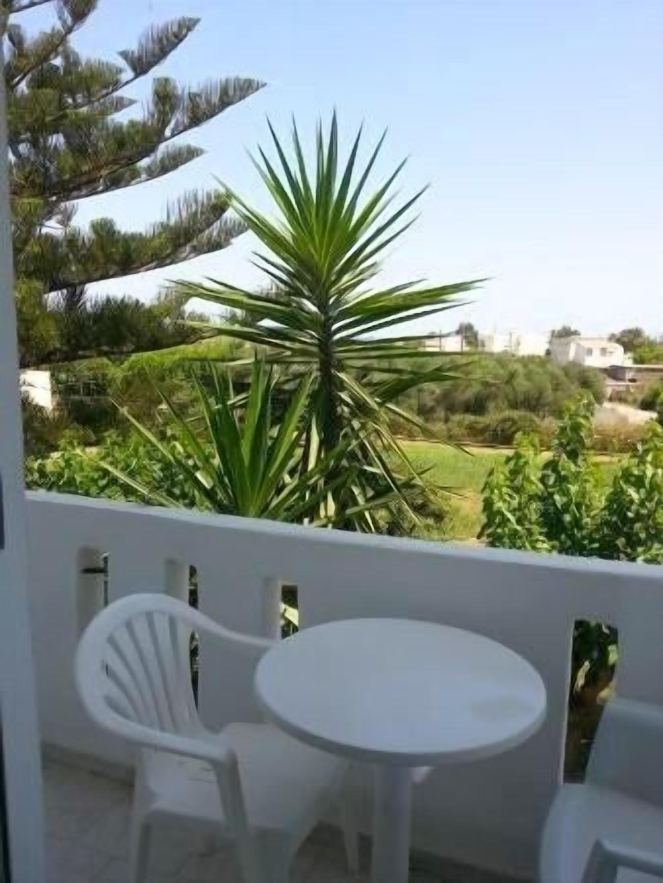 Un balcon avec des chaises et une table en plastique blanc, d’où l’on peut admirer un paysage verdoyant, avec des arbres et des bâtiments.
