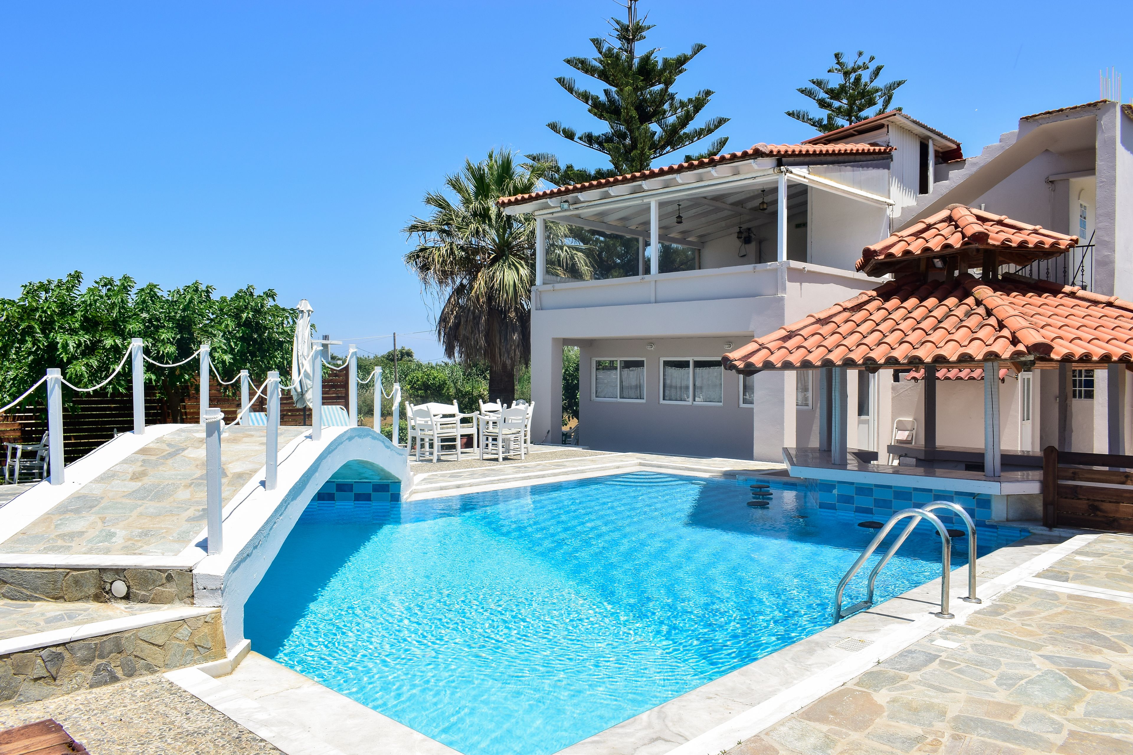 Una piscina con plataforma de piedra, un área de estar cubierta y un edificio de dos pisos con techo de tejas.