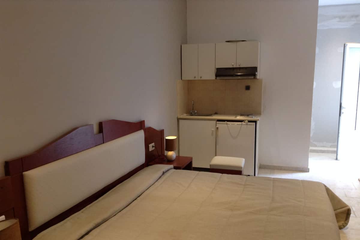A bedroom with a bed, a bedside table, a lamp, a small bench, and a kitchenette with a sink and cabinets.