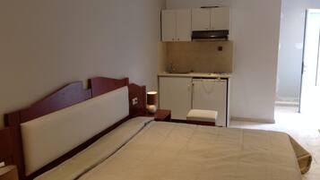 Un dormitorio con cama, mesita de noche, lámpara, pequeño banco y una kitchenette con lavaplatos y armarios.