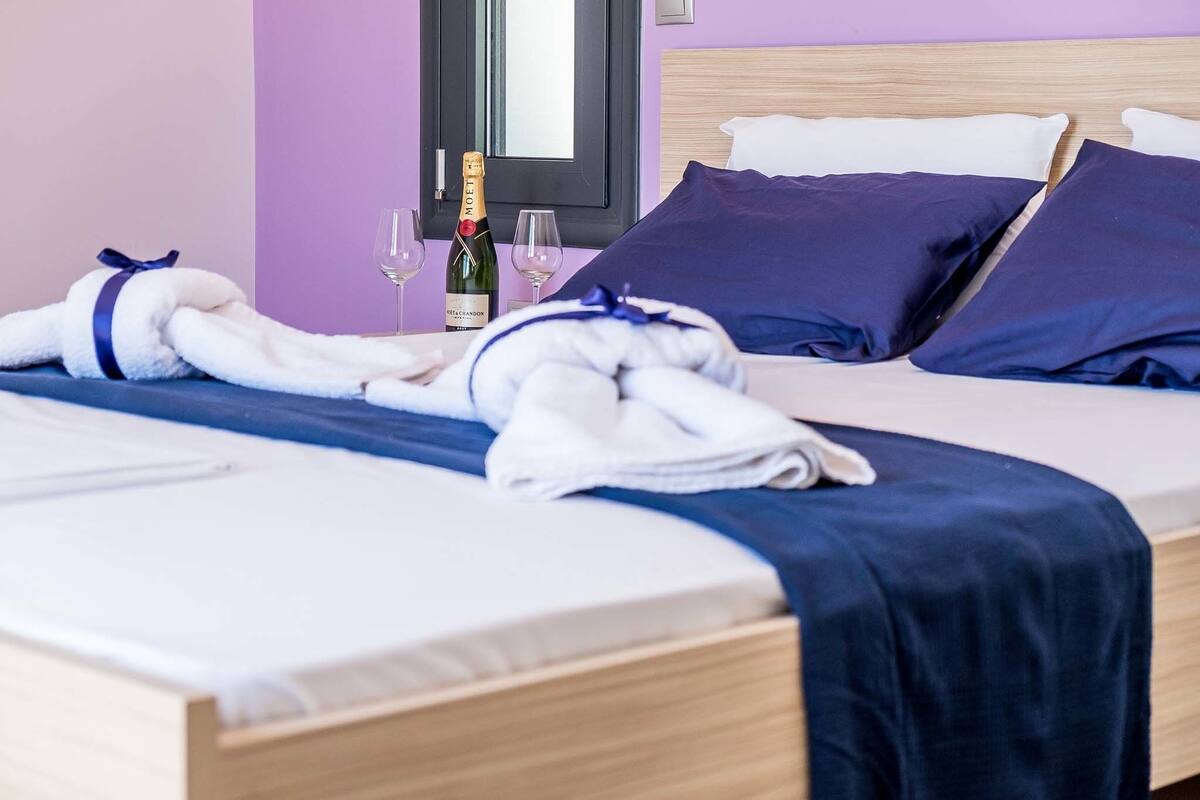 A neatly made bed with a blue bedspread, two pillows, a bottle of champagne, and two glasses on the nightstand.