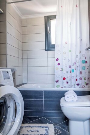 A bathroom with a washing machine, a toilet, and a window with a curtain. - Golden Sun Villas (Lefkada)