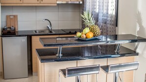 A kitchen with a black countertop, a sink, and a small refrigerator. There is a fruit bowl with a pineapple, an orange, and a pear on the countertop.