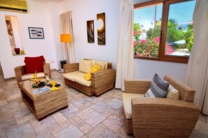 A living room with wicker furniture, a red chair, a yellow cushion, and a view of a garden through a window.