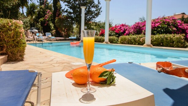 Cenário à beira da piscina com um copo de suco de laranja, duas laranjas e uma espreguiçadeira.