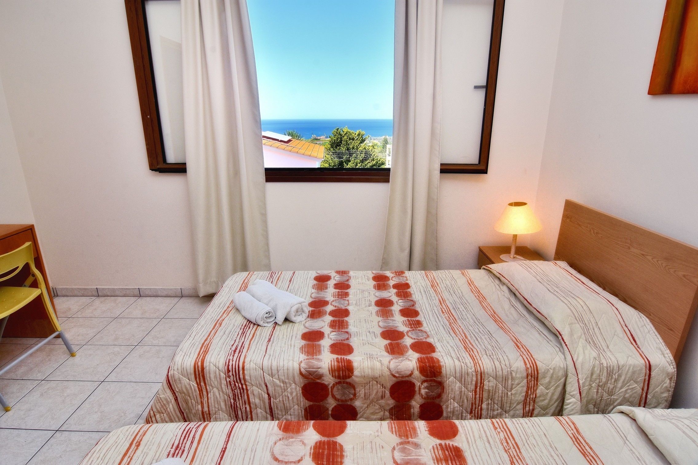 A hotel room with a bed, a nightstand with a lamp, a window with curtains, and a view of the sea.