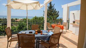 Terraço com mesa posta para dois, rodeado por cadeiras de vime, um guarda-sol branco e vista para o mar.